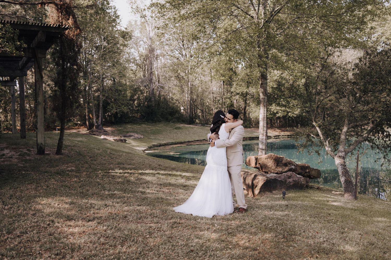 Wedding couple shares intimate moment by turquoise pond surrounded by trees.