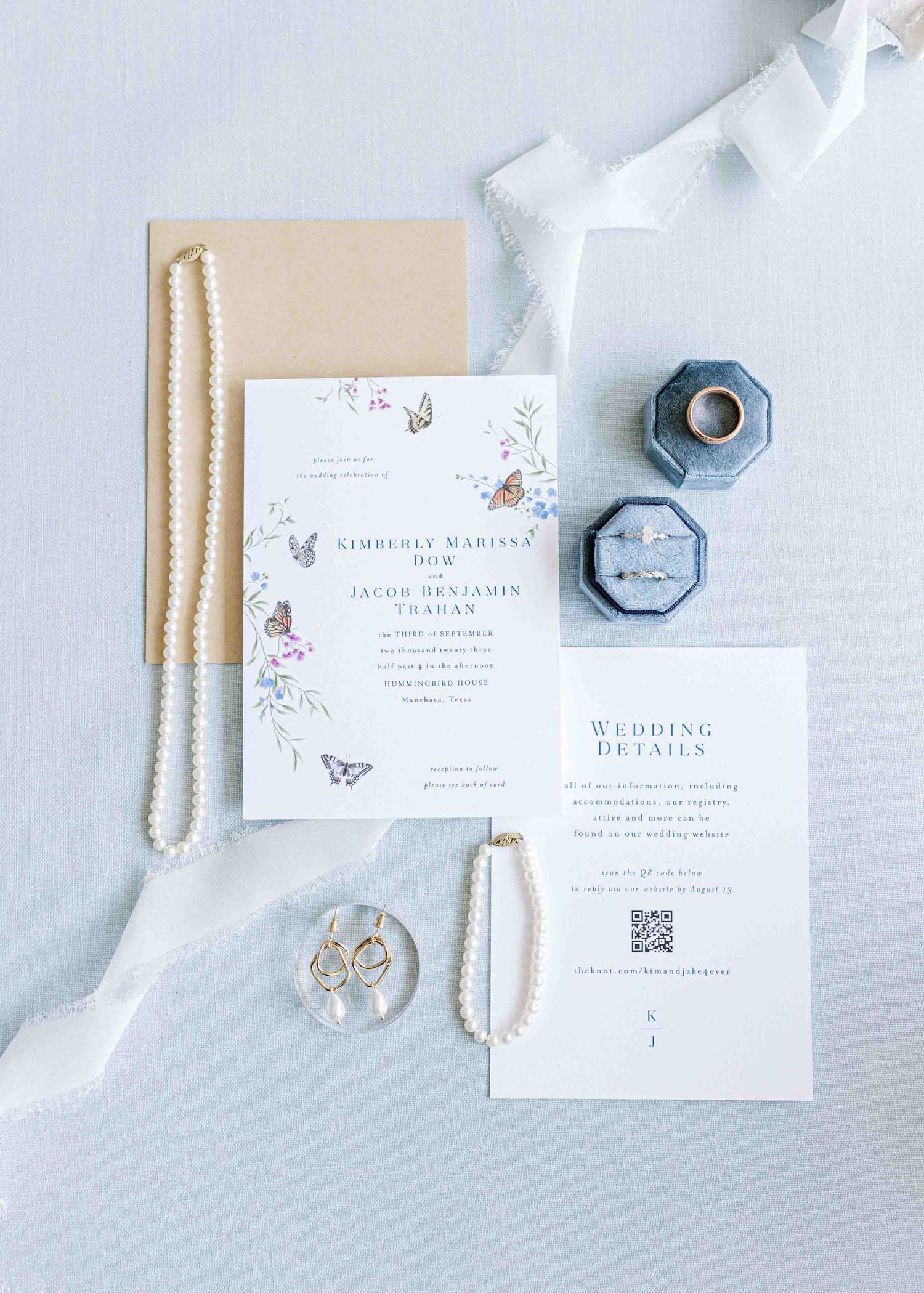 Elegant wedding invitation suite with floral details, pearls, and rings on light gray background.