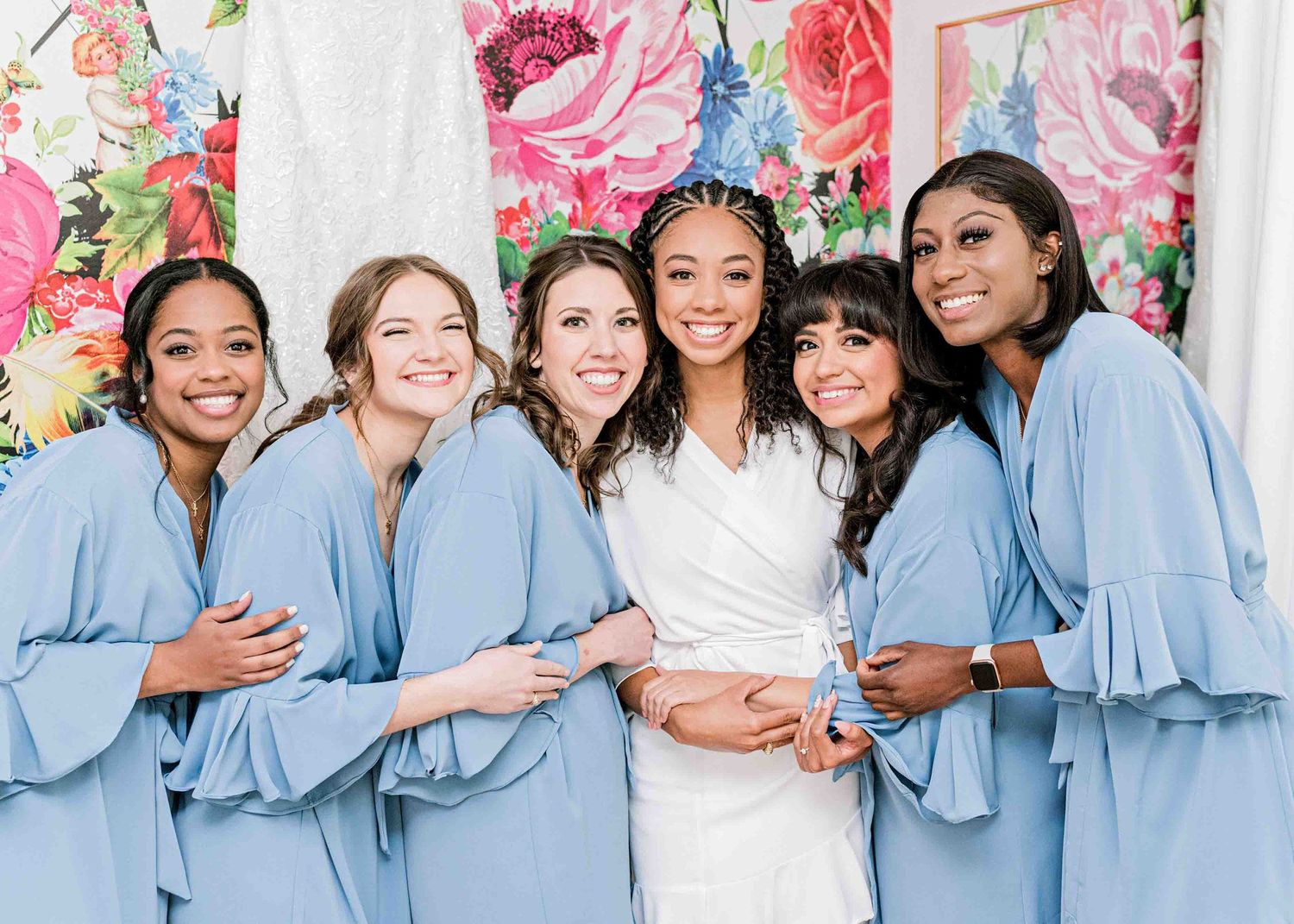 Wedding party group photo with bridesmaids in light blue robes against floral backdrop.