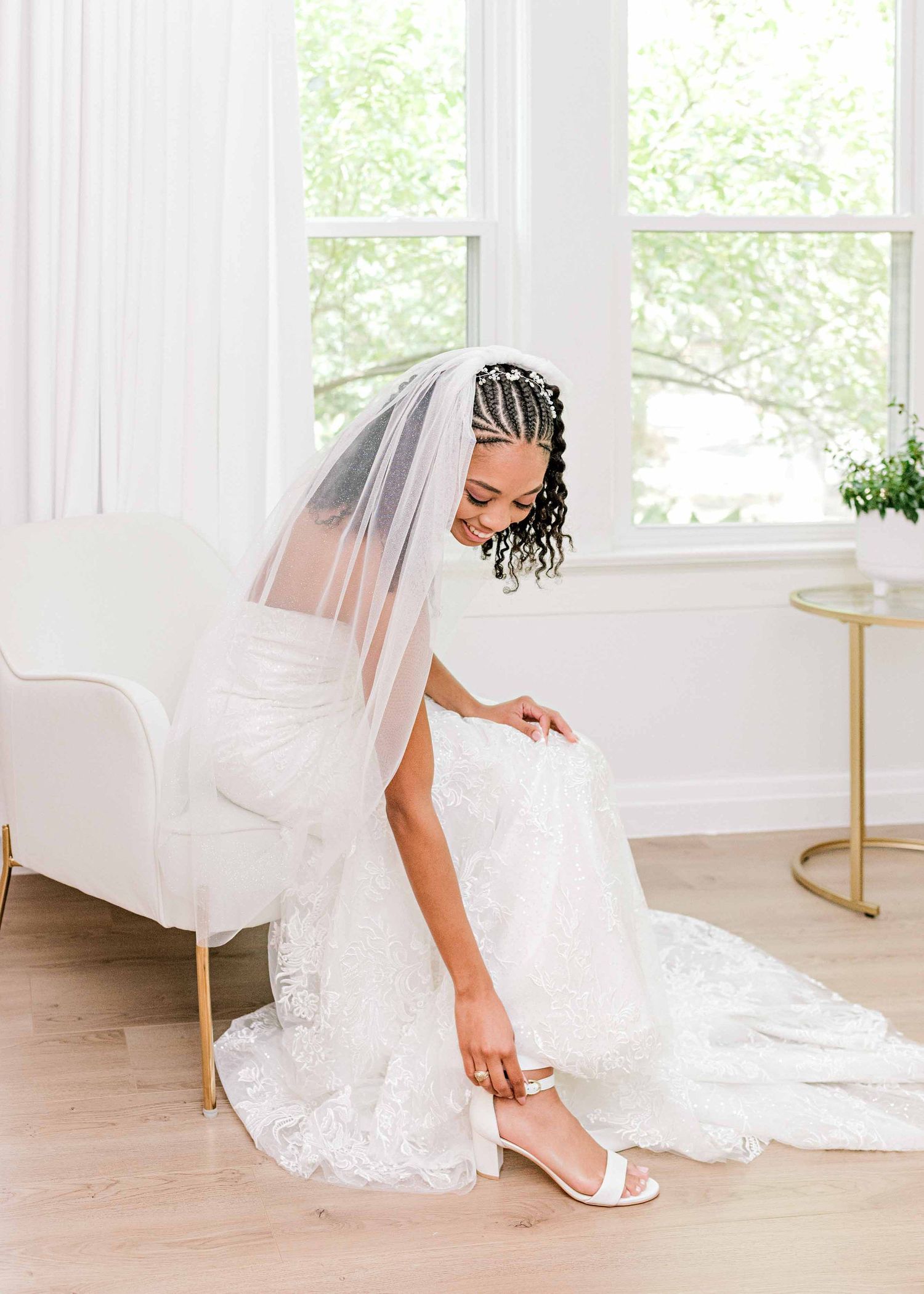 Bride in white lace gown adjusting shoes in bright, modern getting ready room.