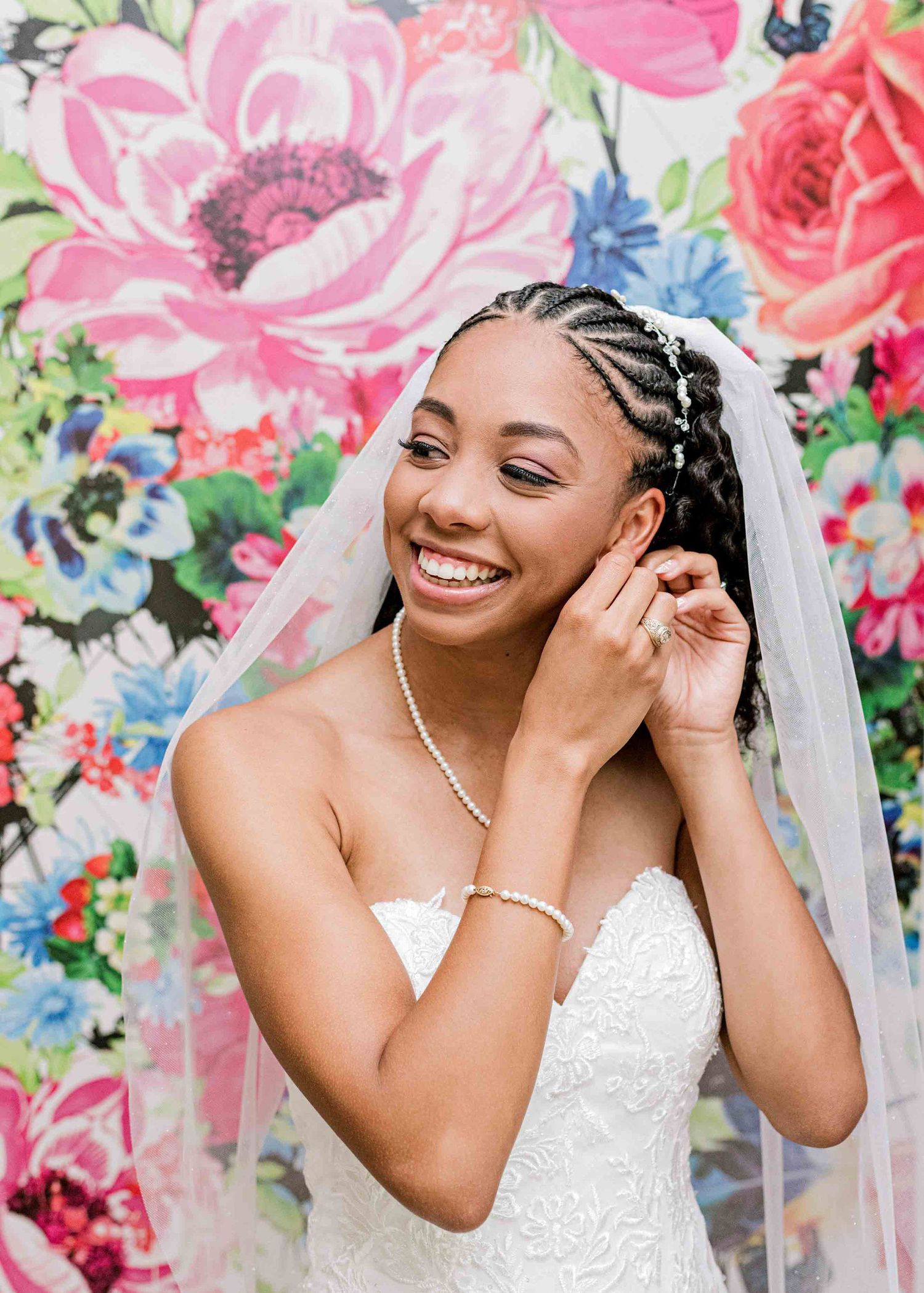 Beautiful bride in a lace gown putting on earrings against a floral wallpaper.