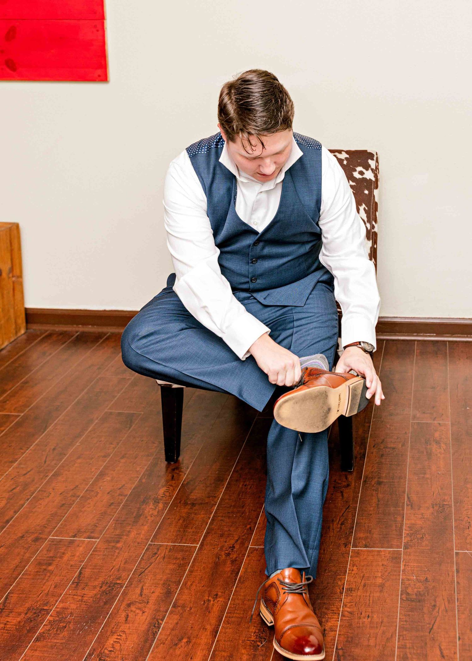 Groom in navy vest sitting on chair while adjusting his shoe.