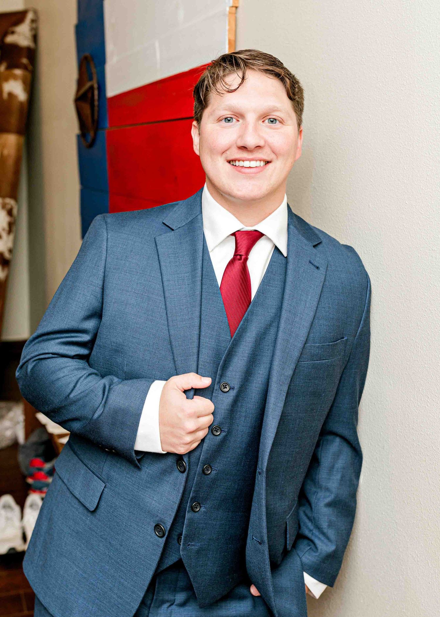 Groom in blue suit and red tie standing against red and blue wall.