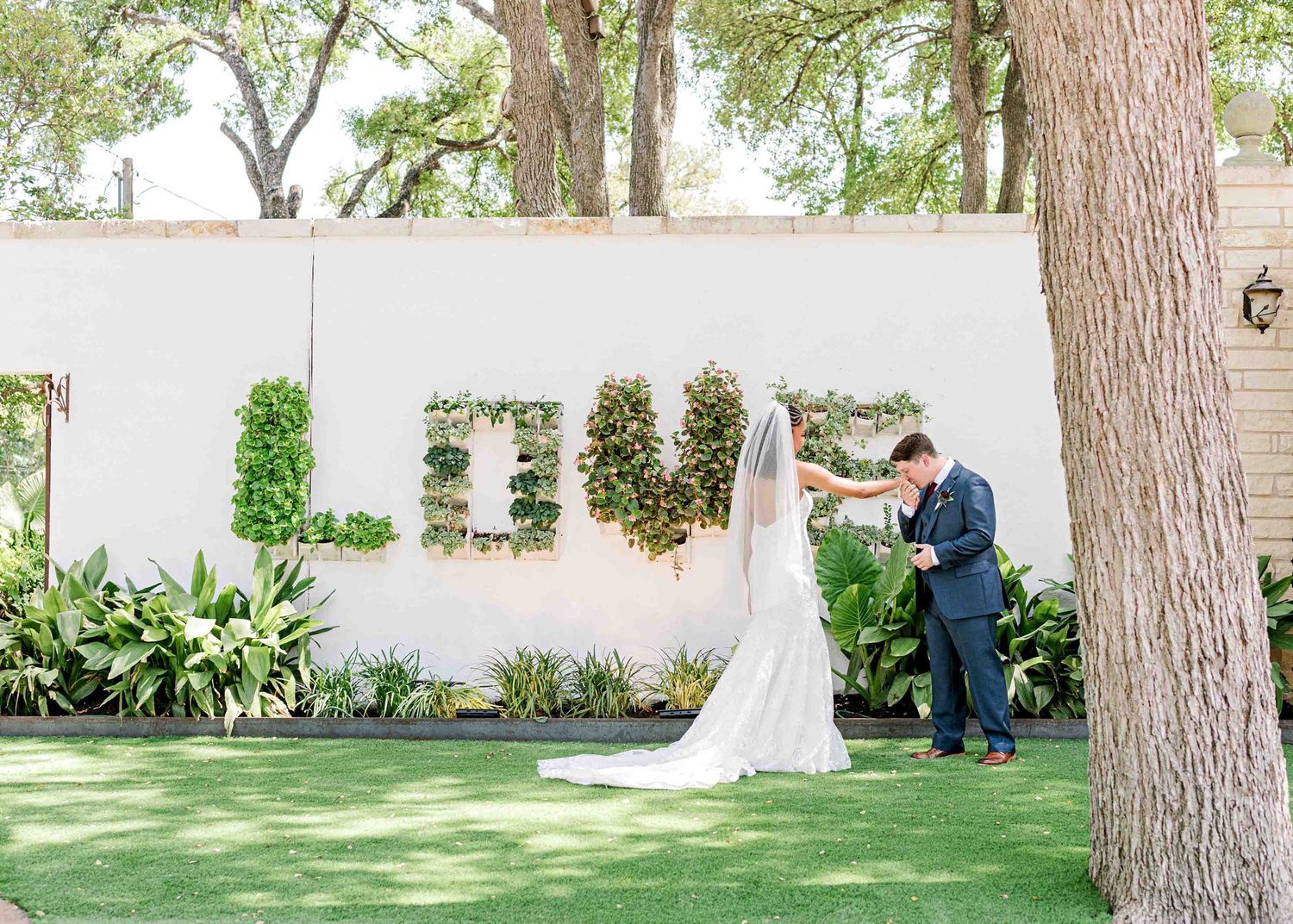 Elegant outdoor wedding ceremony setup with floral LOVE sign against a white wall surrounded by trees.