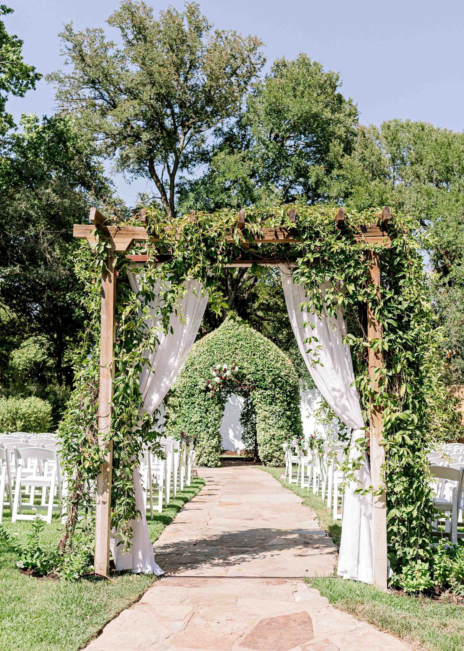 Romantic wedding aisle with wooden pergola, white drapes, and lined ceremony chairs.