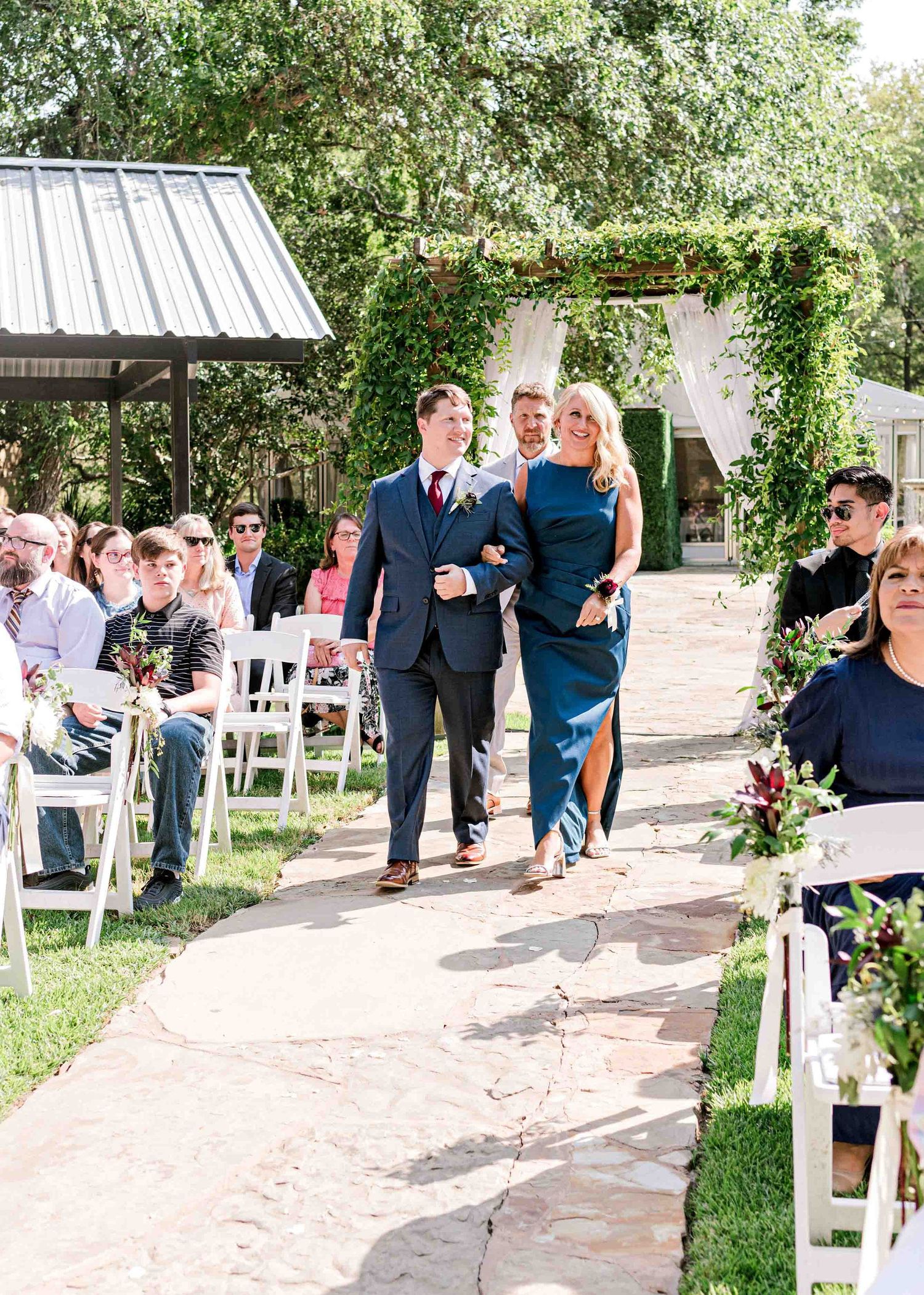 A church wedding processional walking down an outdoor stone pathway lined with white chairs and floral decor.