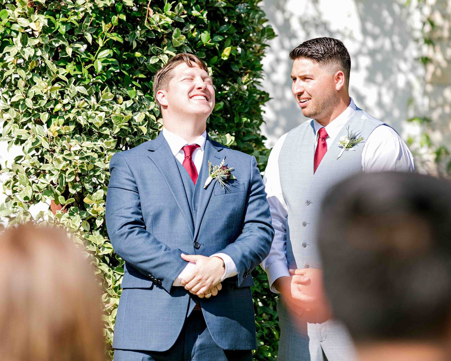 Two attendants share a moment at an outdoor wedding ceremony under green foliage.