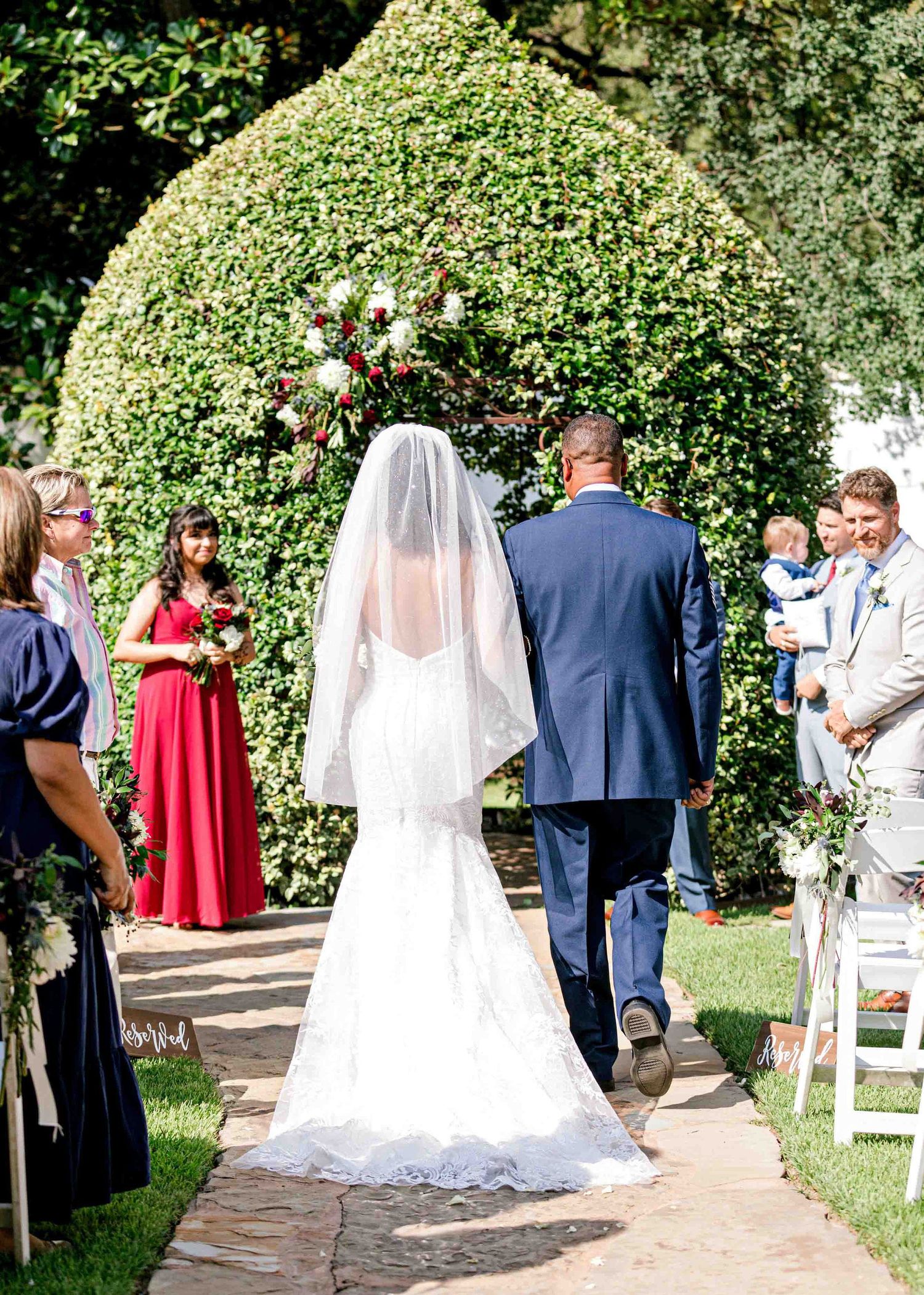 A wedding ceremony in progress under a lush green garden arch with guests seated on white chairs.