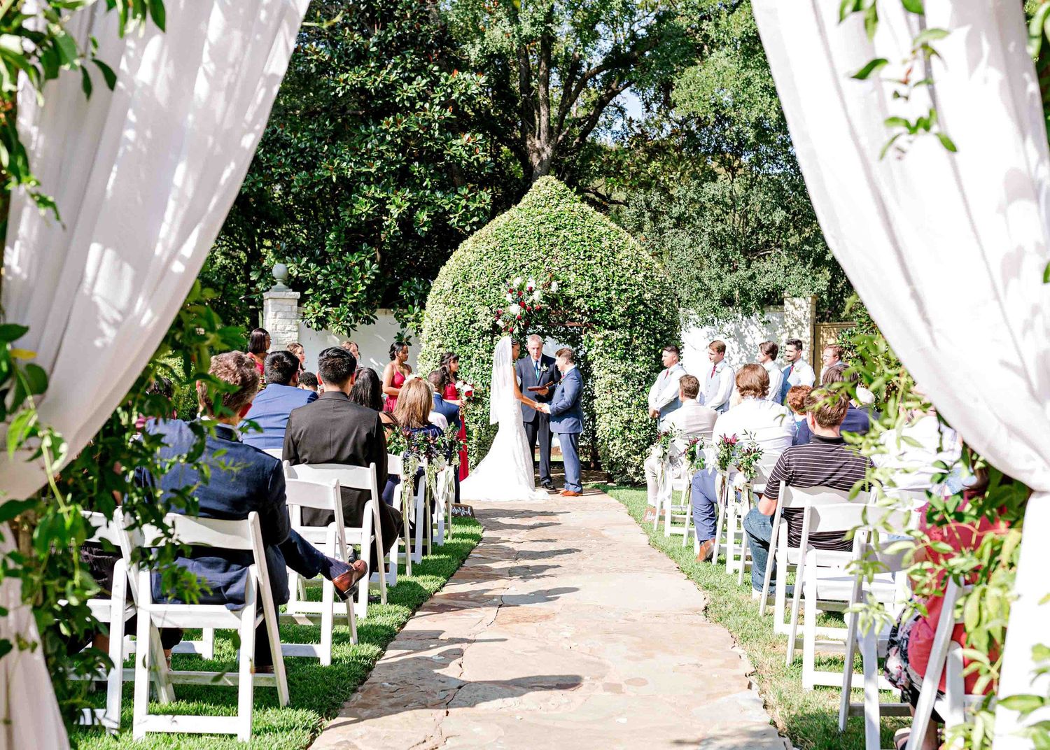 A garden wedding ceremony with white drapes and chairs lining a stone pathway under green arches.