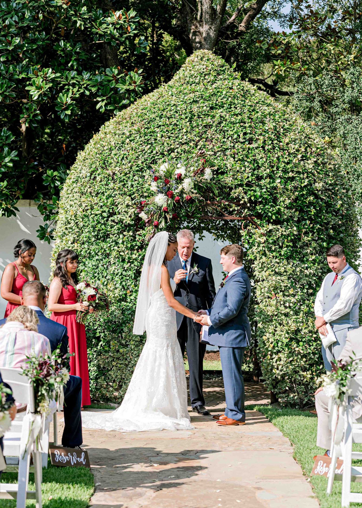 A wedding ceremony in progress under a manicured garden arch surrounded by guests in white chairs.