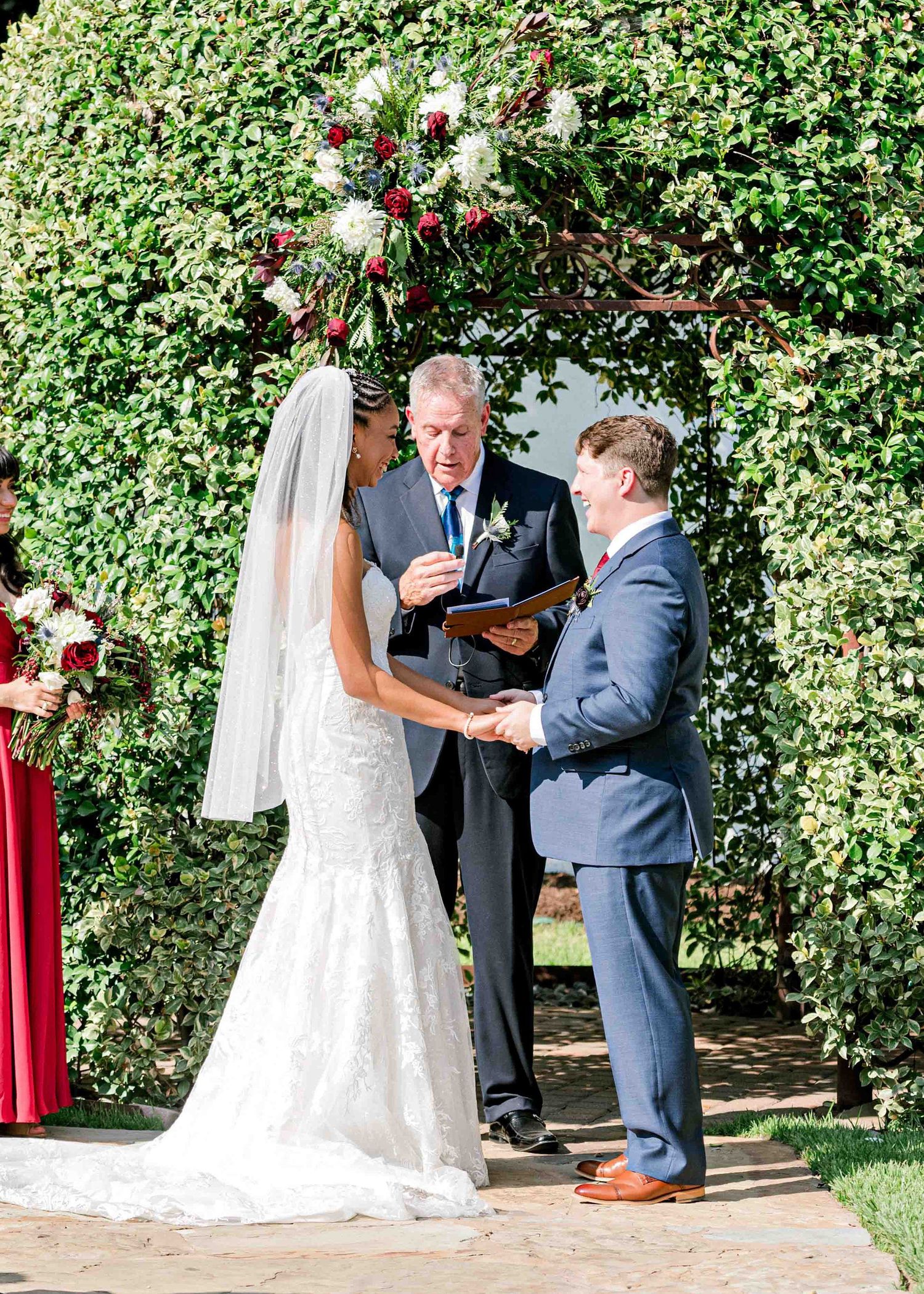 Wedding ceremony taking place under a beautiful floral arch with red roses and lush greenery.