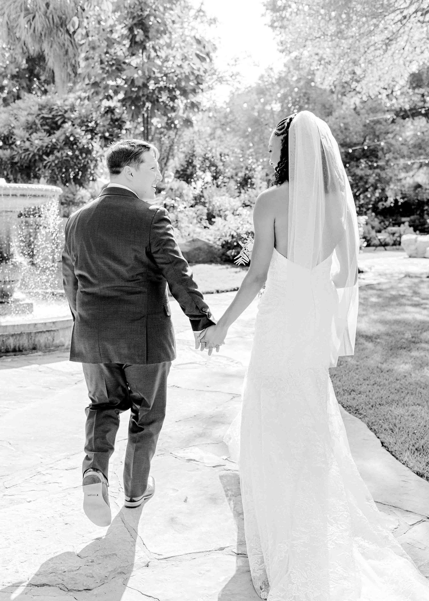 Black and white photo of newlyweds walking hand in hand along a garden path.