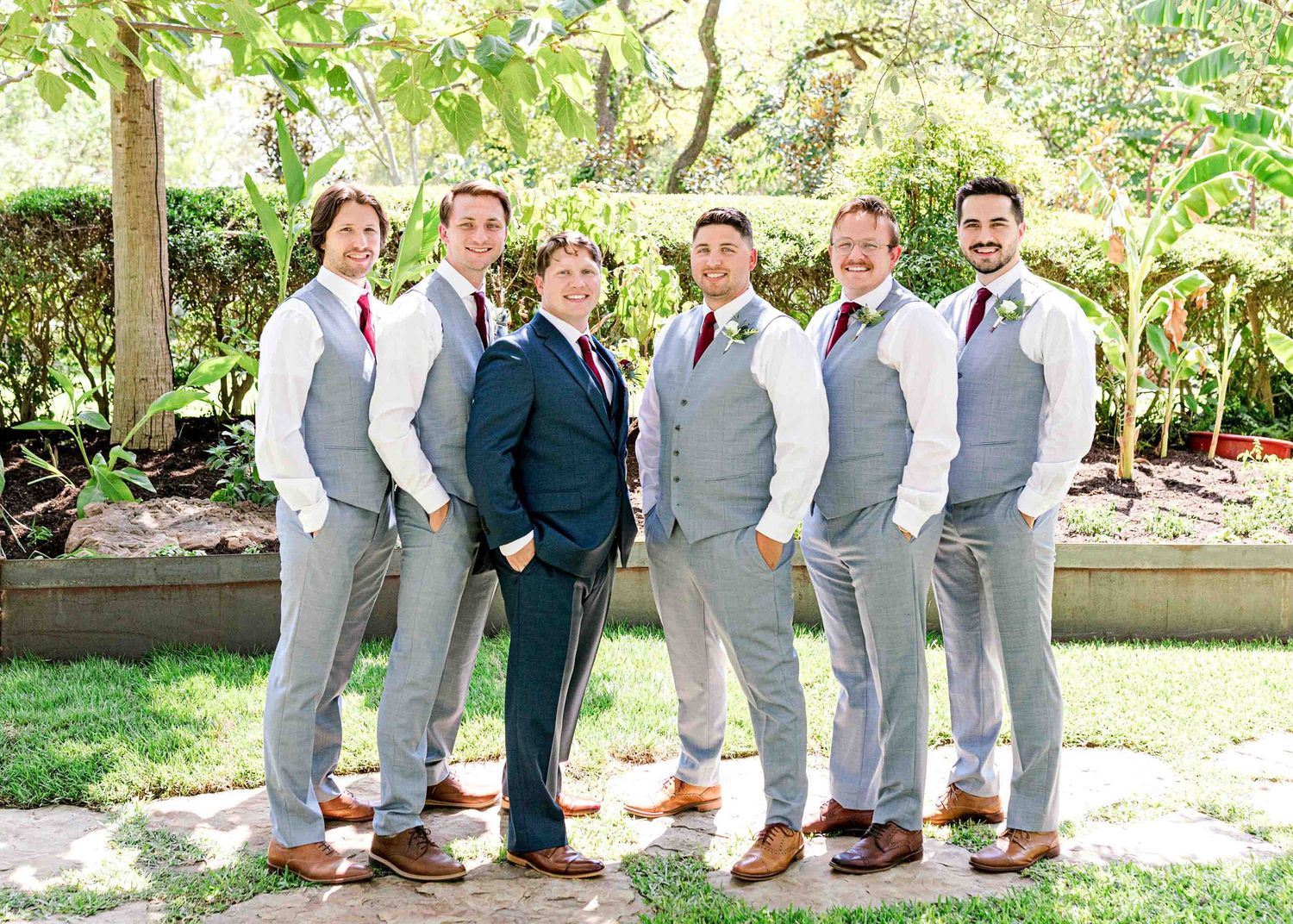 Wedding party in coordinated grey suits and navy blue standing together in a garden.