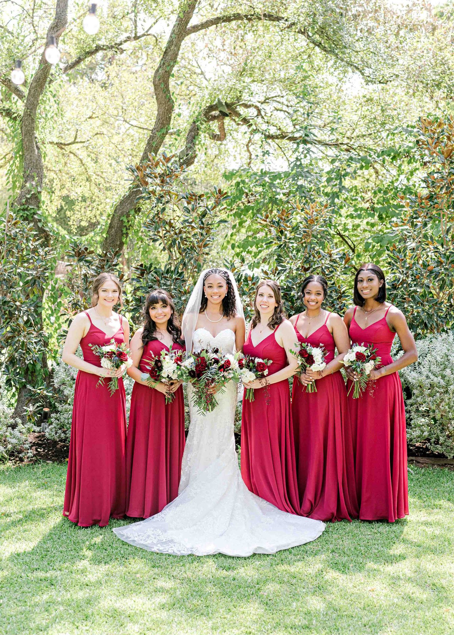 Wedding party photo with bridesmaids in flowing red dresses holding bouquets in a garden setting.
