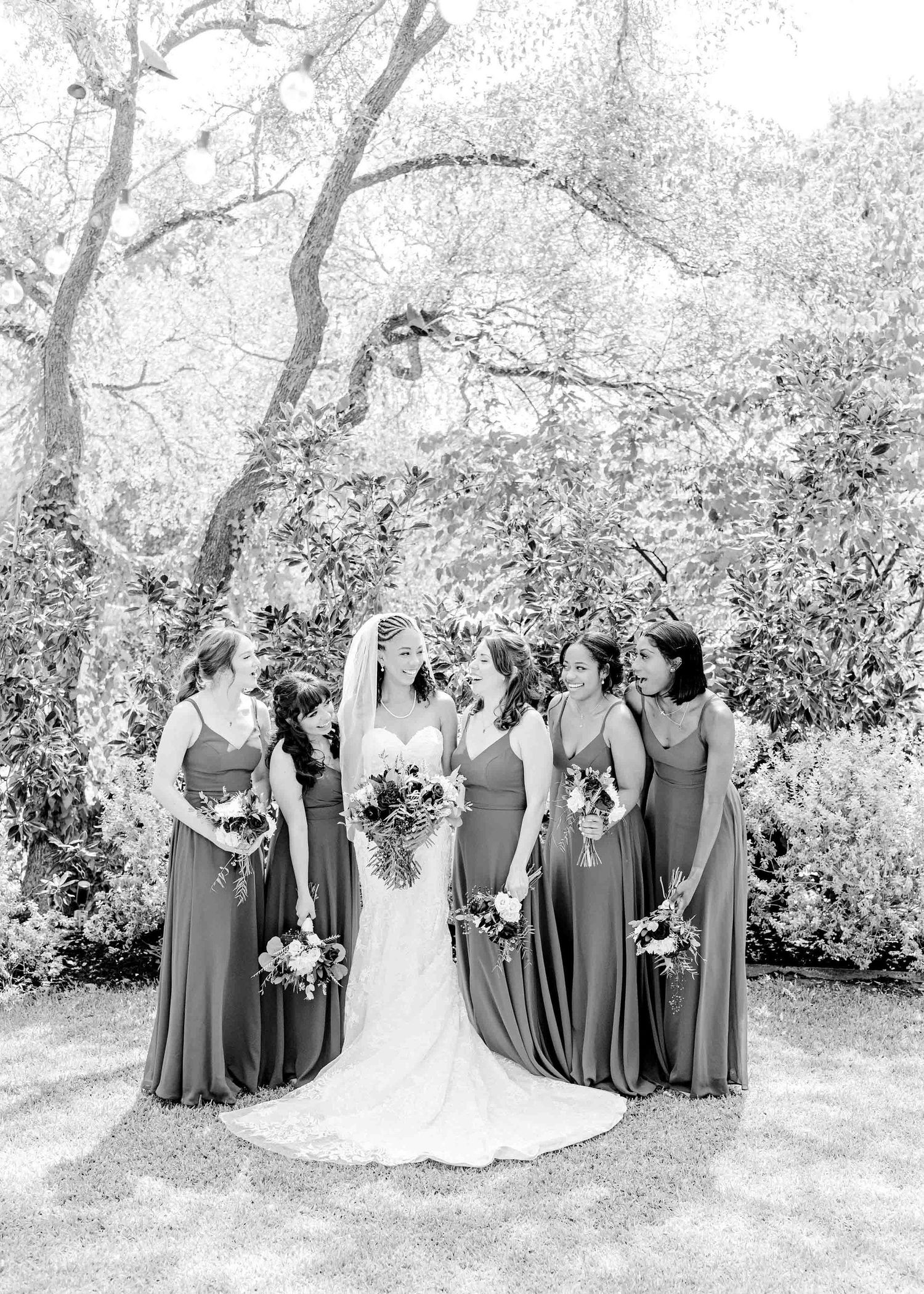 Black and white photo of a wedding party with bridesmaids in elegant long dresses surrounded by lush garden scenery.