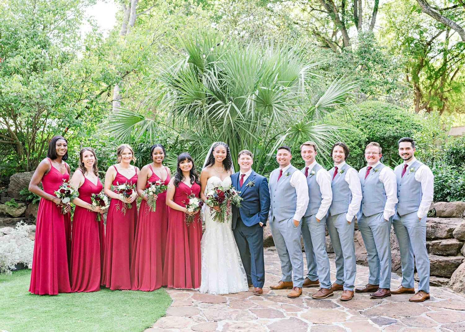 Wedding party in red bridesmaid dresses and light gray suits poses on stone steps in a beautiful garden setting.