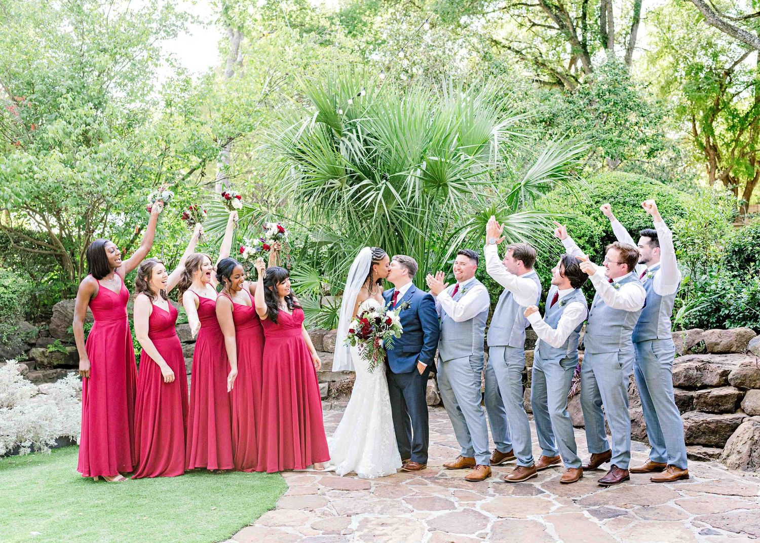 Wedding party cheering with arms raised on garden stairs with bridesmaids in red and groomsmen in gray.
