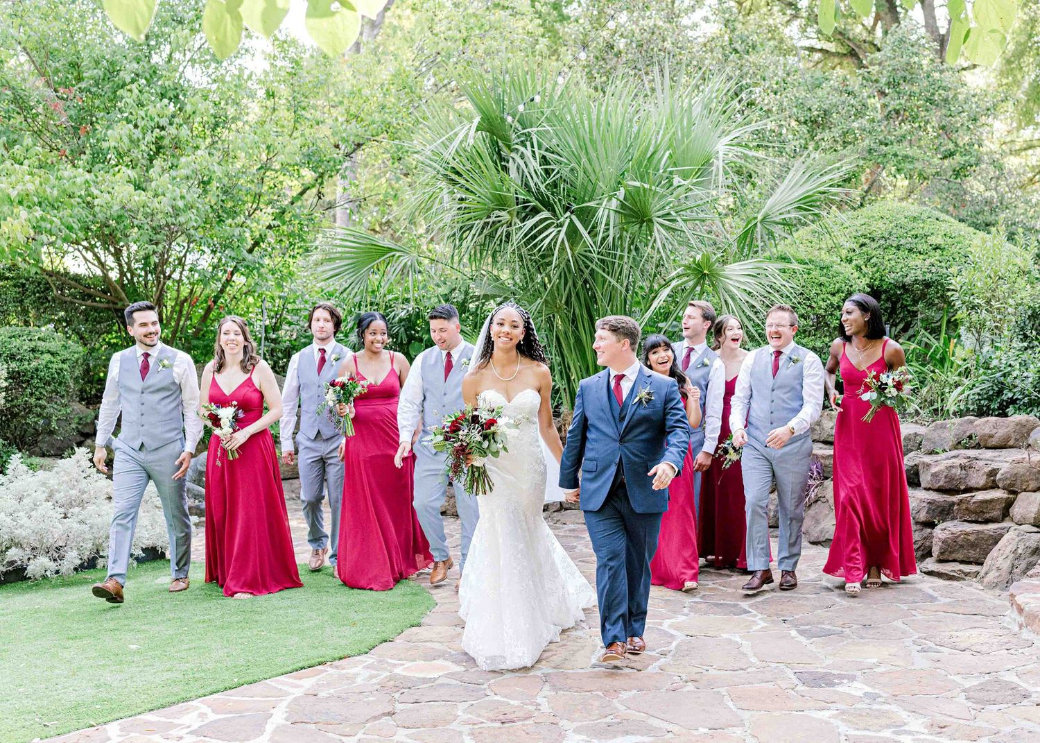 Wedding party poses casually on garden stone steps with bridesmaids in red gowns and groomsmen in gray suits.