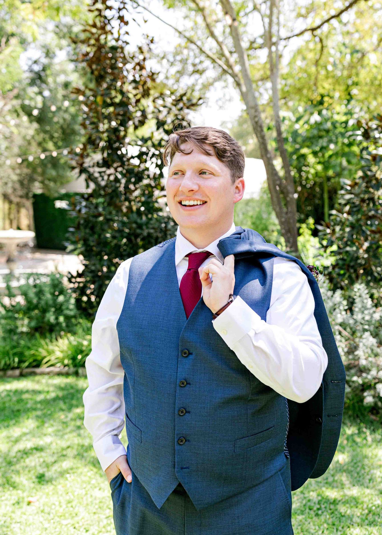 Groom in navy vest and red tie smiling in a sunlit garden setting.