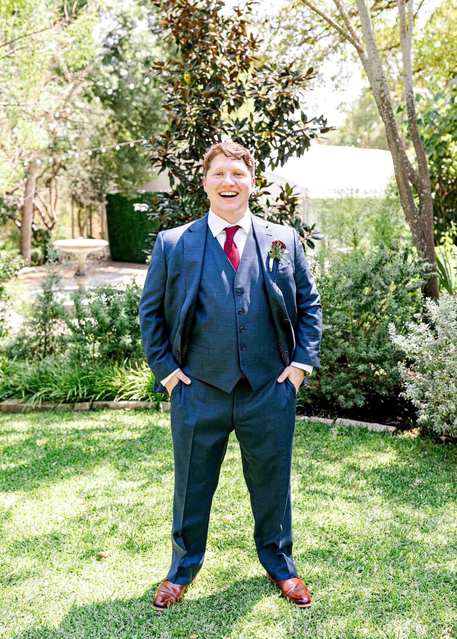 A wedding guest in a navy three piece suit poses on a sunny lawn with trees in the background.