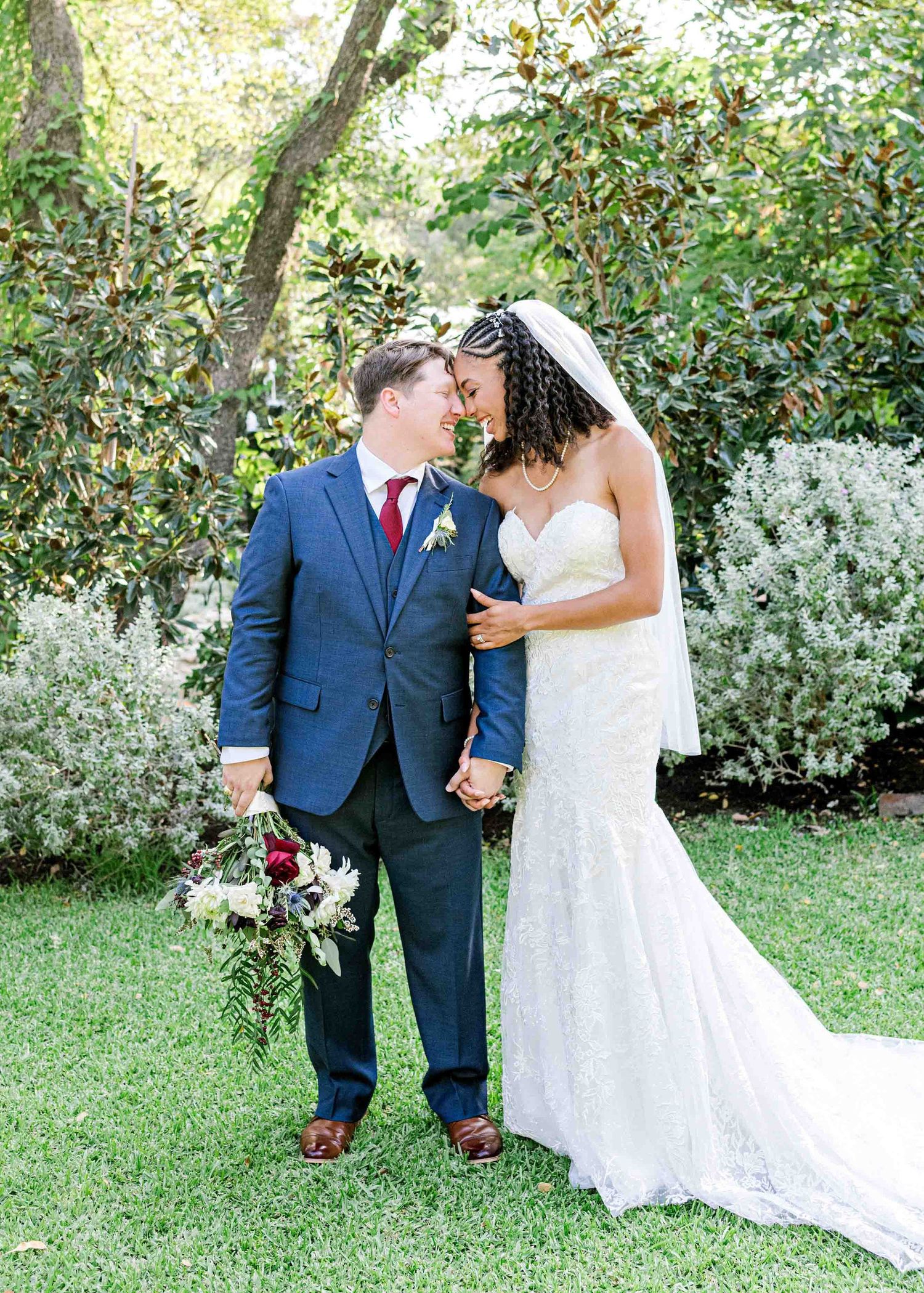 A bride in white lace gown and groom in navy suit share a moment in a garden setting.
