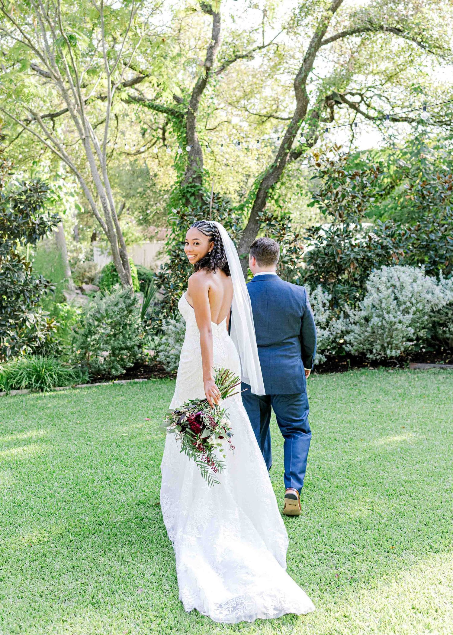 Bride in flowing white dress holds bouquet while standing in a sunlit garden.