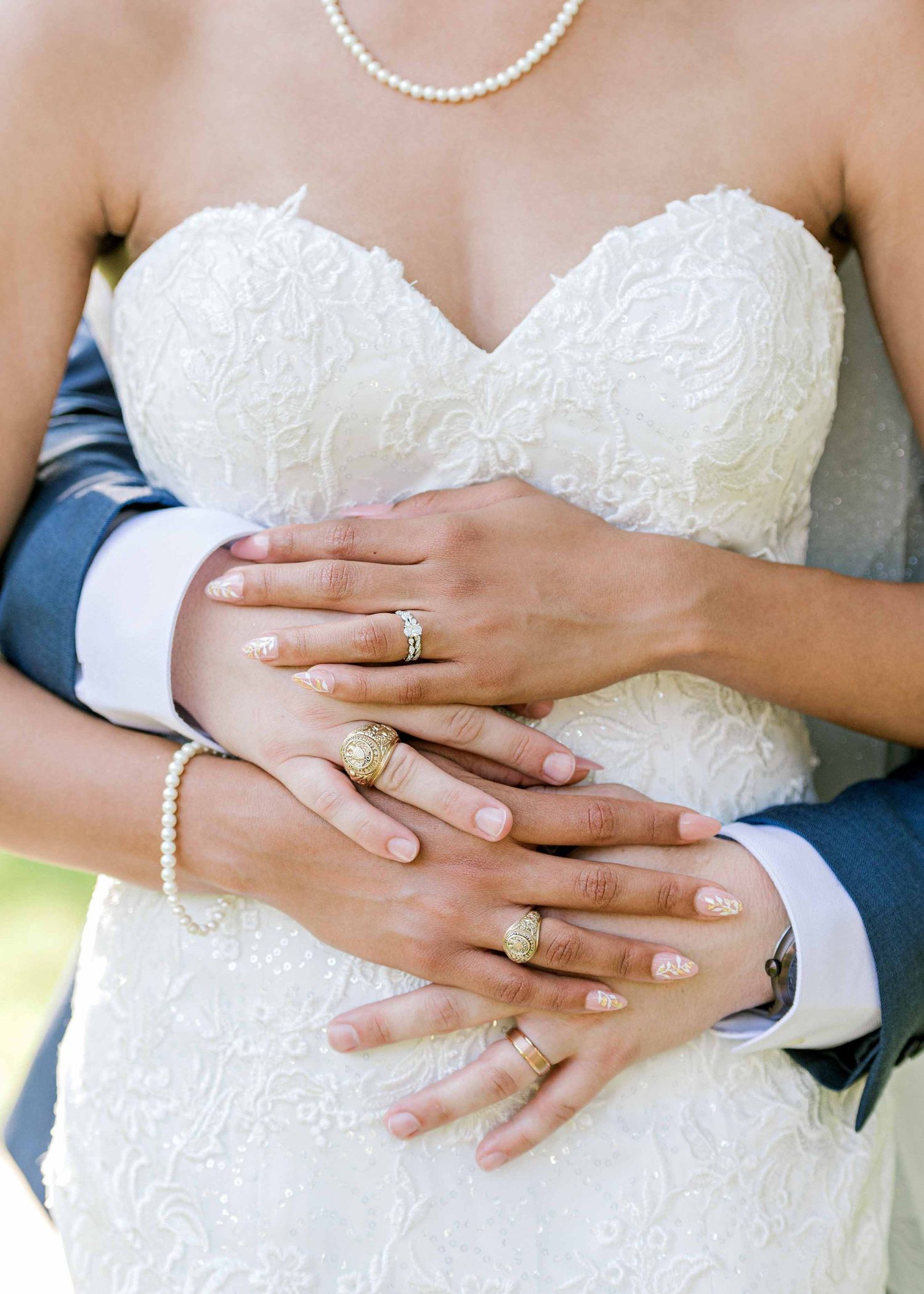 Close-up of hands with wedding rings against white lace dress and navy suit.