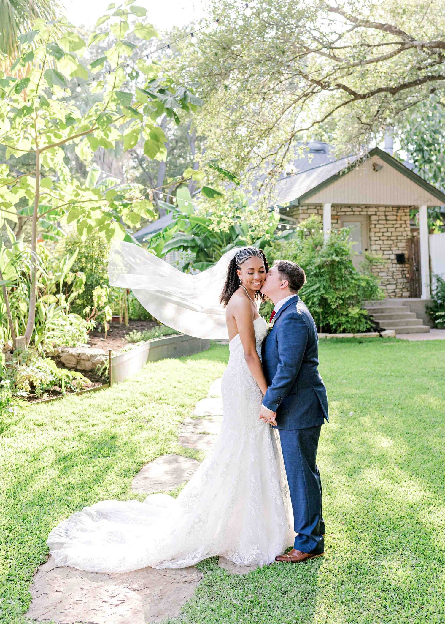 Romantic garden wedding portrait with flowing veil and stone cottage in background.