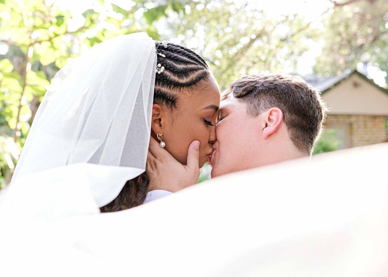 Intimate wedding kiss captured through flowing white veil in garden setting.
