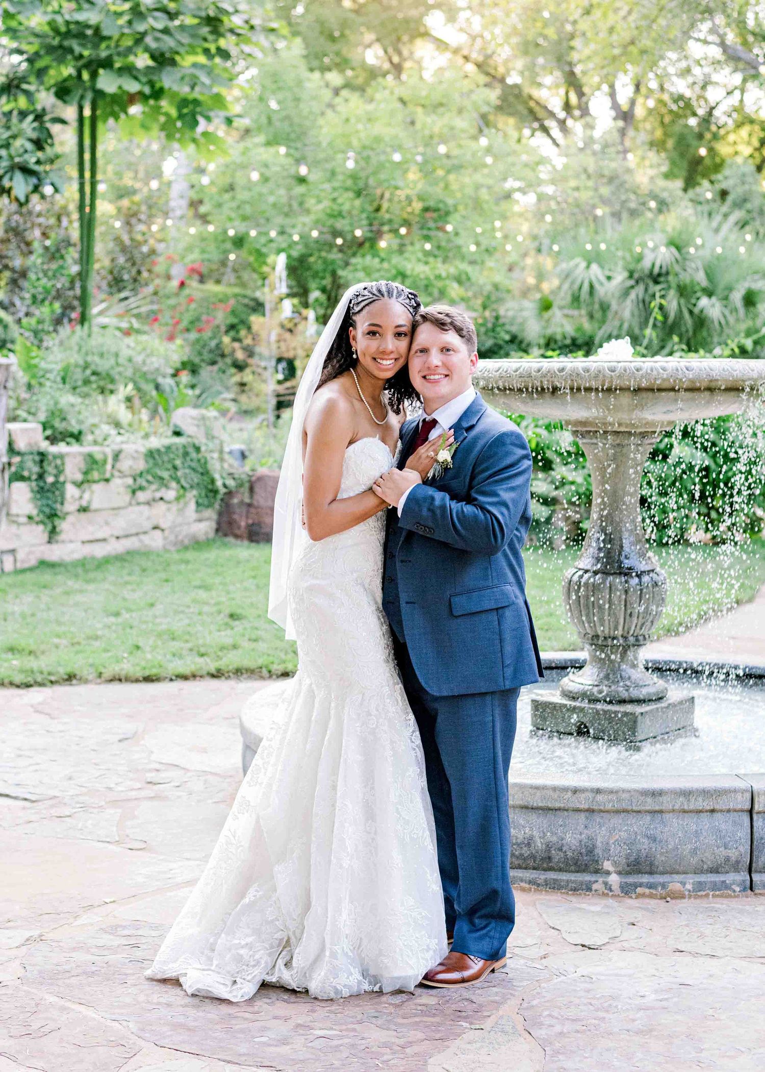 Elegant wedding portrait by stone fountain in lush garden setting.