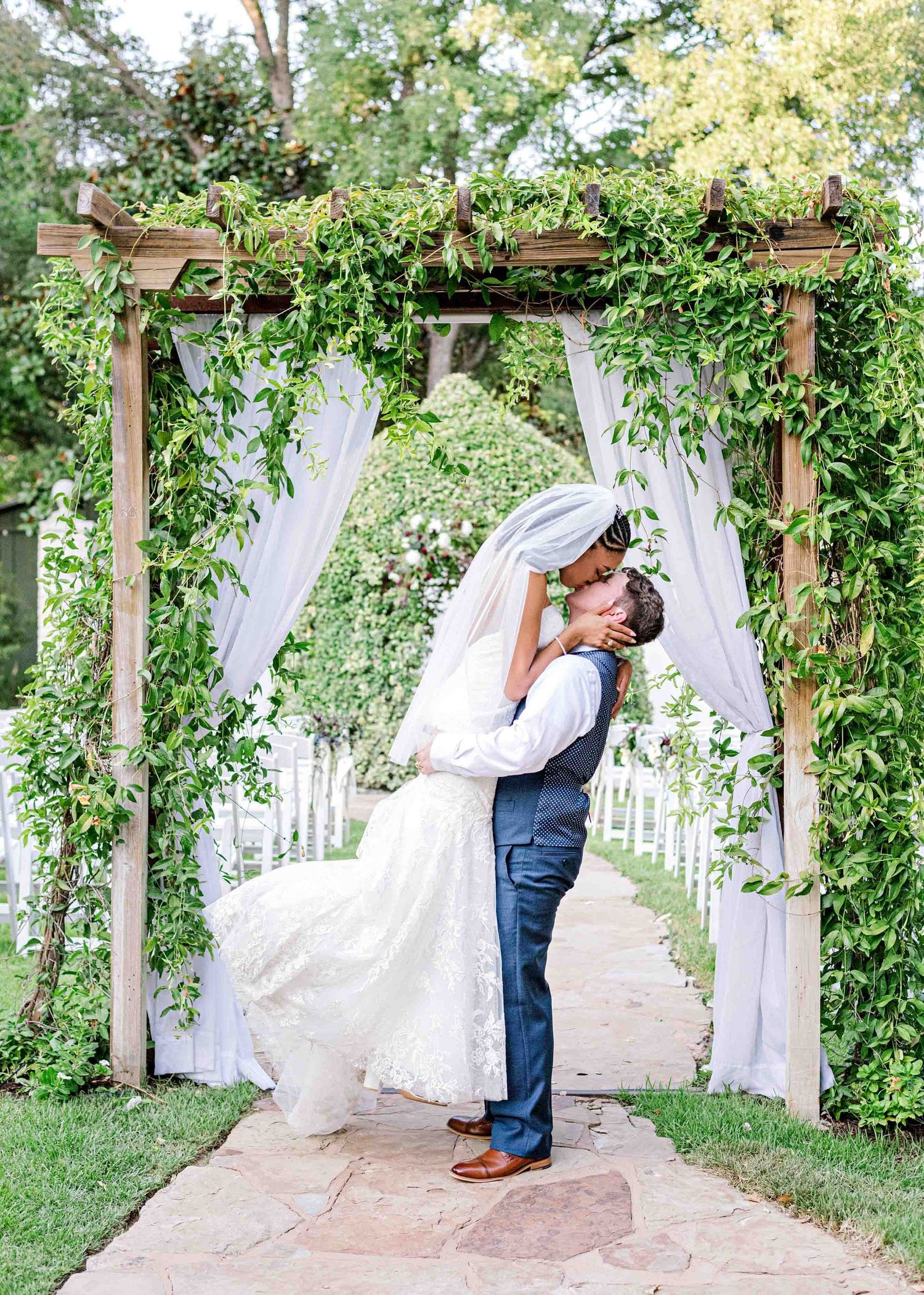 Romantic embrace under vine-covered wooden arch with flowing white drapes.