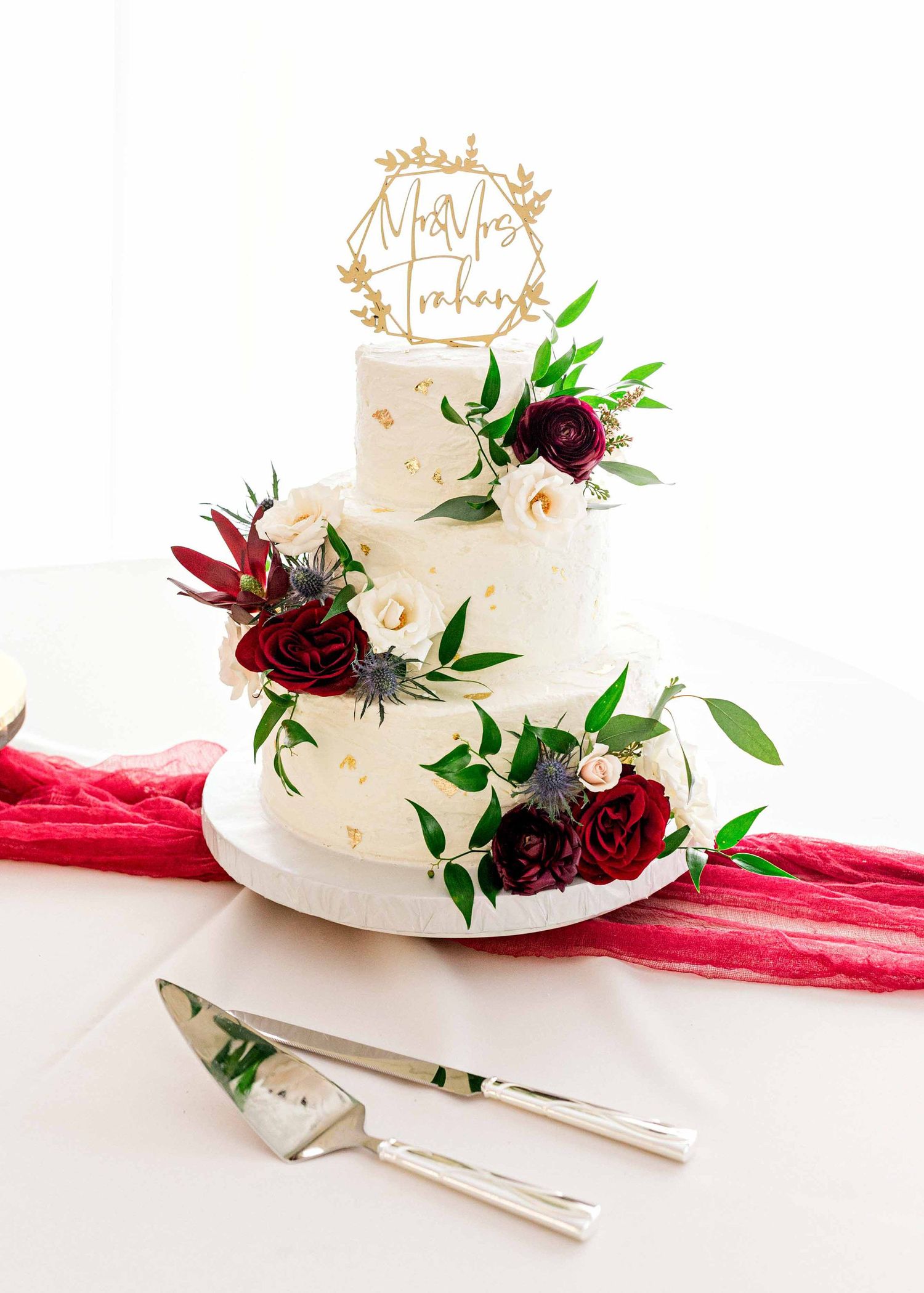 Elegant white wedding cake decorated with burgundy flowers and gold geometric topper.