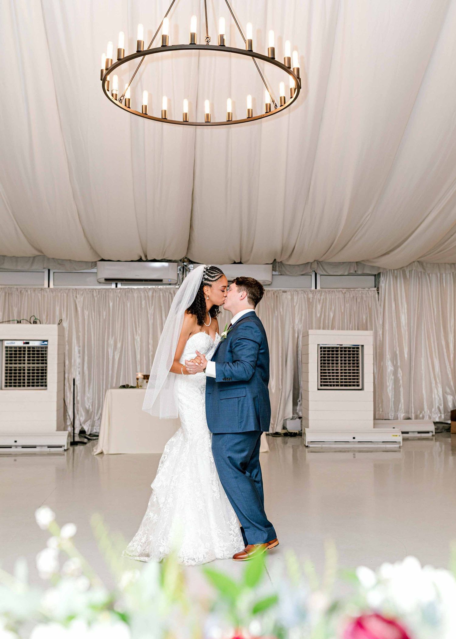 Couple dancing beneath a circular chandelier in an elegantly draped white reception space.
