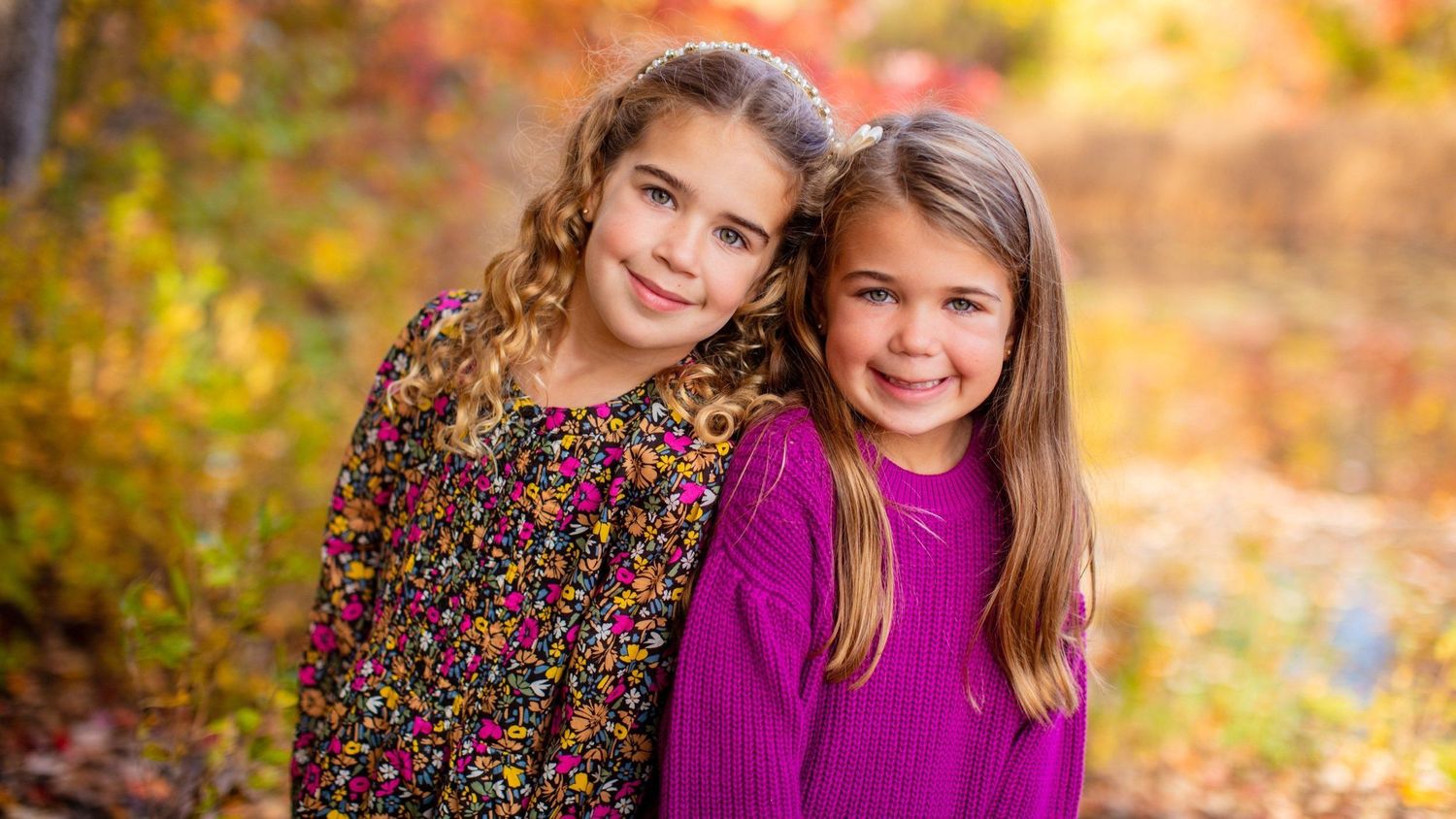 Two smiling girls pose together outdoors among vibrant autumn foliage, one in floral dress, one in purple sweater. for Family Photos Shoot