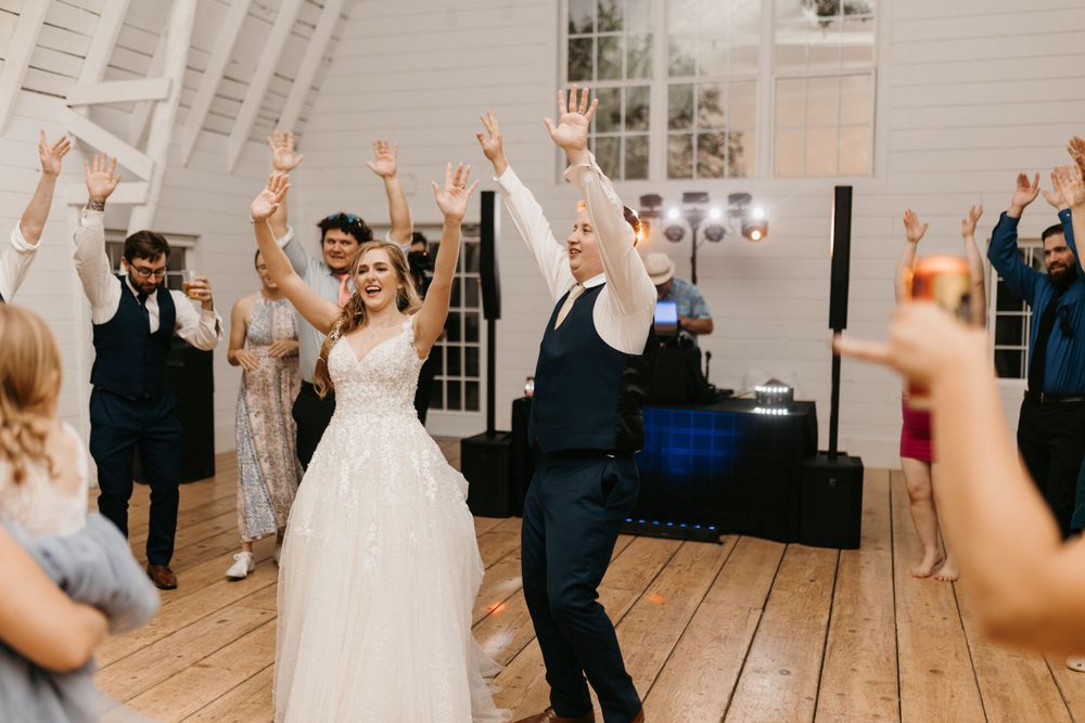 Bride and groom raise hands with cheering wedding guests during energetic reception dance celebration.