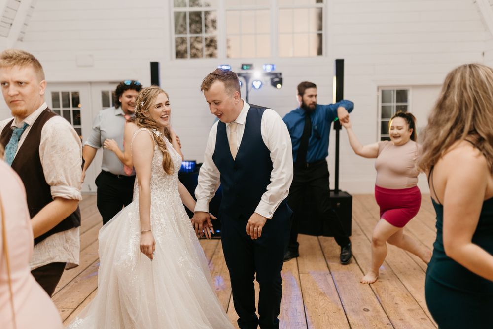 Bride and groom dancing joyfully with wedding guests on a wooden dance floor at their reception.