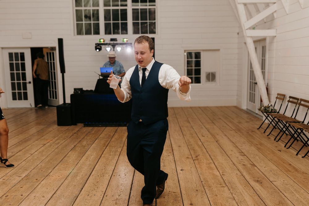 Groomsman in navy suit performing solo dance moves on wooden floor at wedding reception.