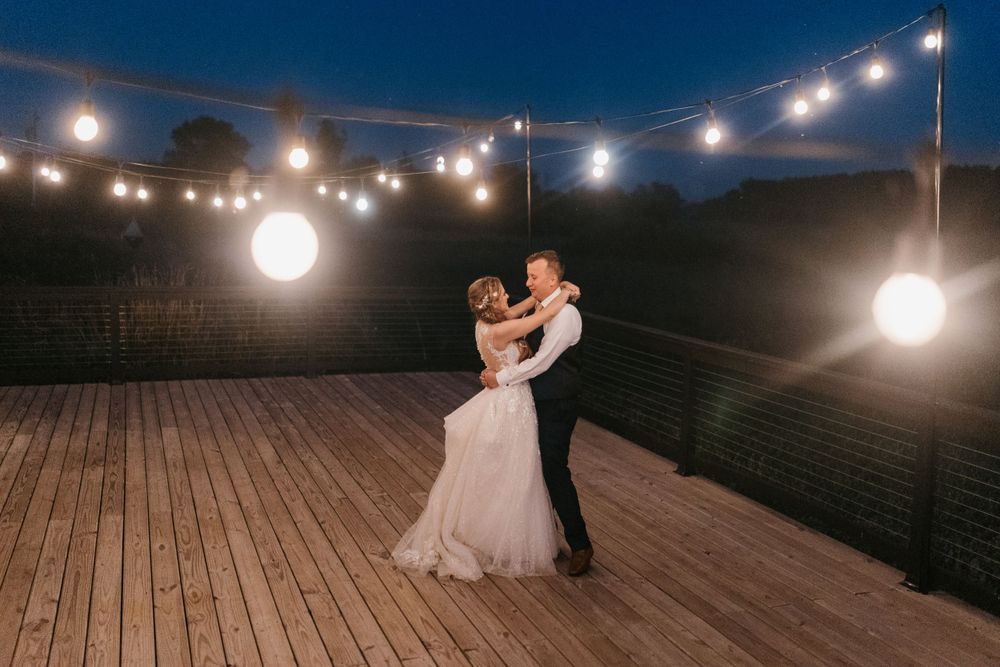 Newlyweds share their first dance under glowing string lights on an outdoor wooden deck at night.