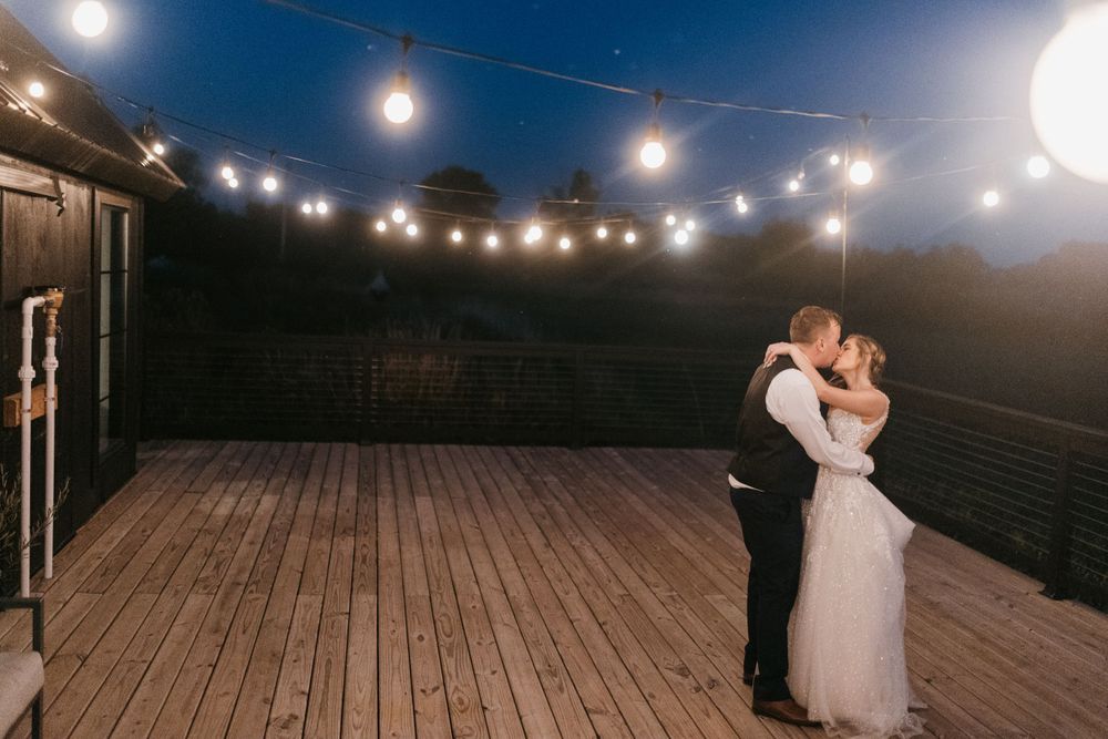 Bride and groom share a romantic first kiss under string lights on a wooden deck at twilight.