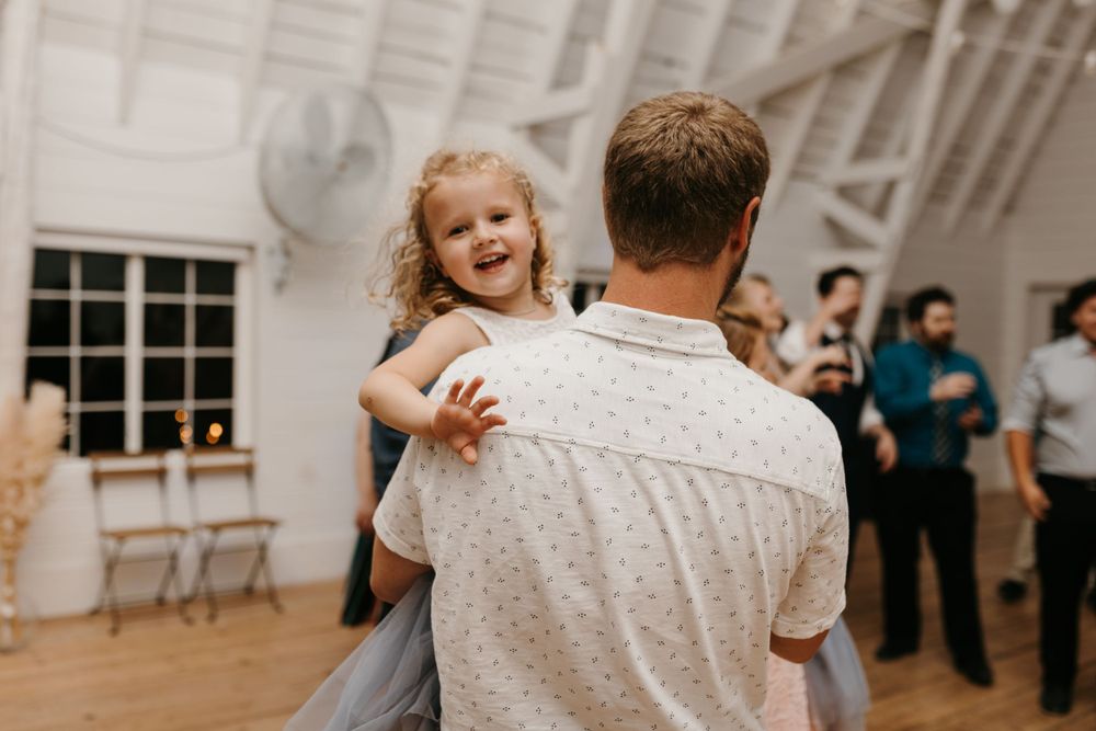 Young curly-haired flower girl smiling while being held and danced with by a man at wedding reception.