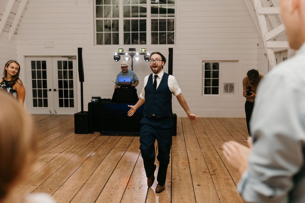 Man in navy vest and glasses dancing energetically on wooden dance floor at wedding reception.