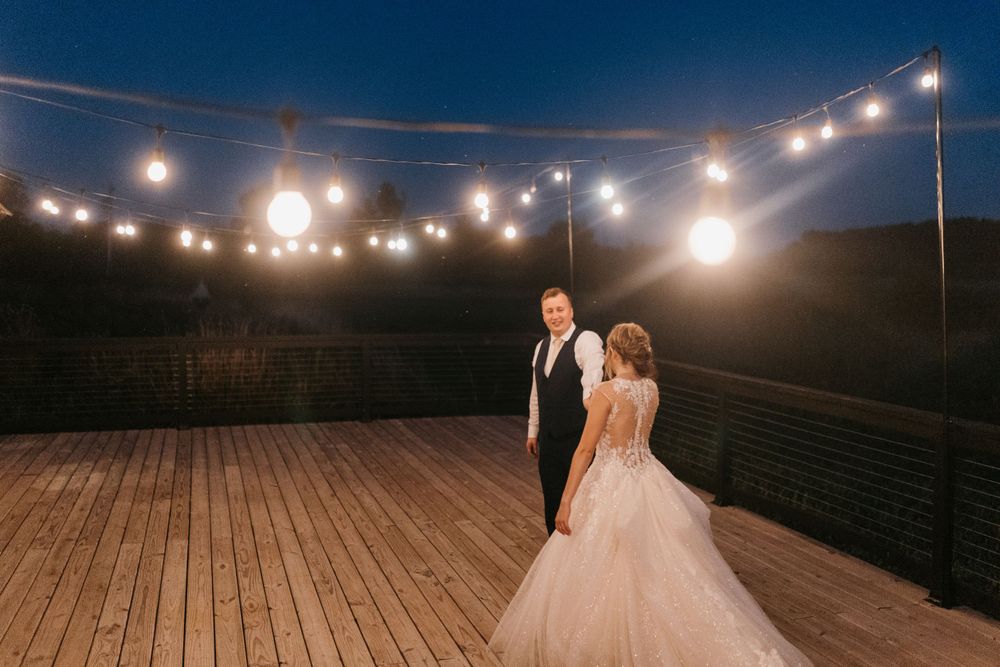 Bride and groom share a tender moment under warm string lights on a wooden deck at dusk.