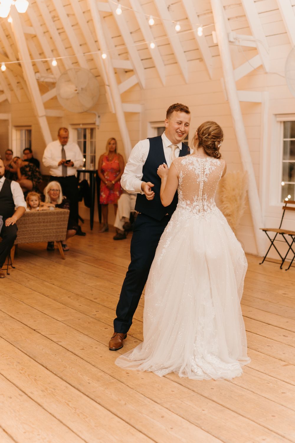 Smiling groom dances with bride in a beautiful lace-back wedding gown in a rustic barn reception hall.