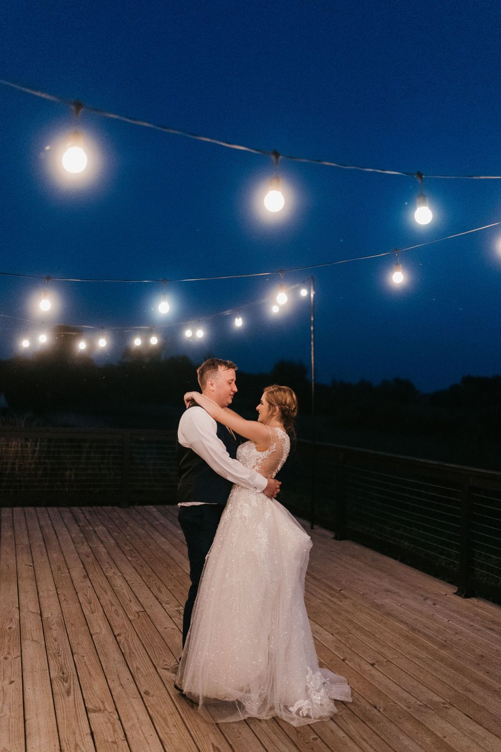 Bride and groom share romantic dance under glowing string lights on outdoor deck at twilight.