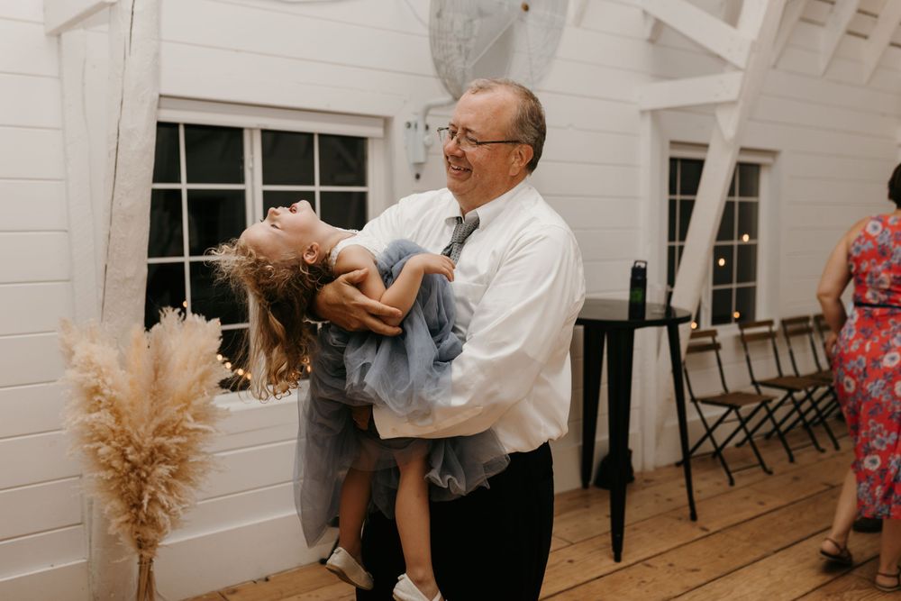 Older man in white shirt and tie joyfully lifting and dancing with a young girl at wedding reception.