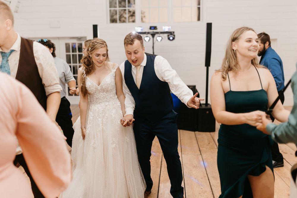 Smiling bride in lace gown and groom in navy vest dancing hand-in-hand with guests at wedding reception.