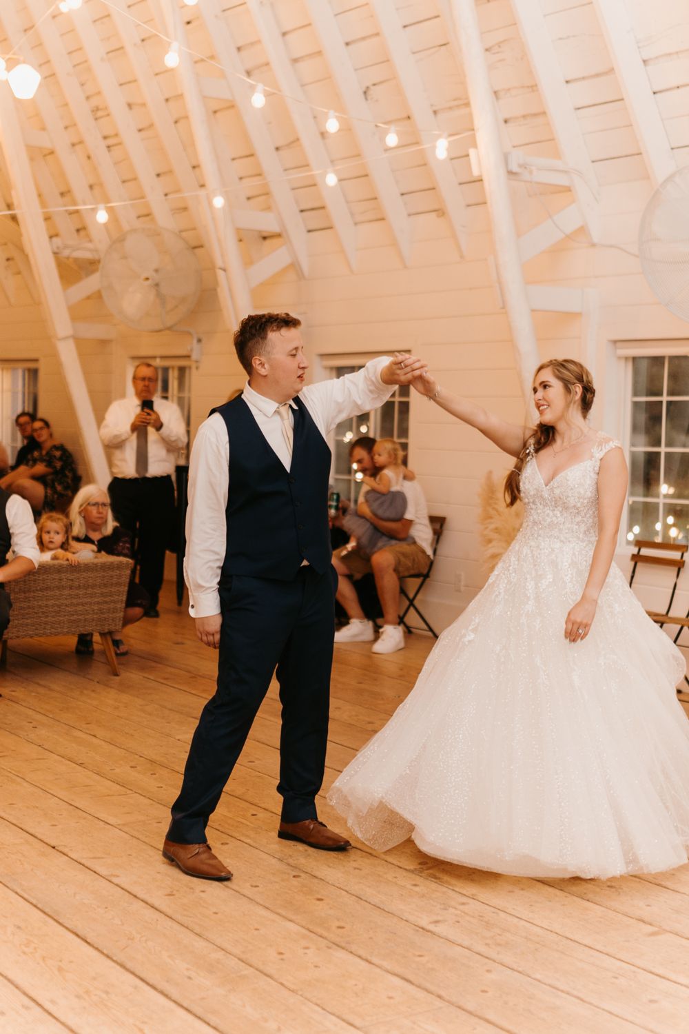 Groom twirls bride in a stunning lace gown during their first dance in a warmly lit barn venue.