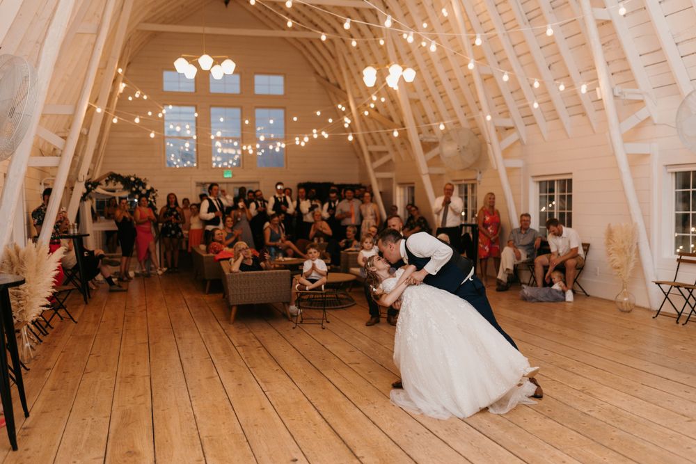 Bride and groom share a romantic dip during their first dance in a barn wedding venue with guests watching.
