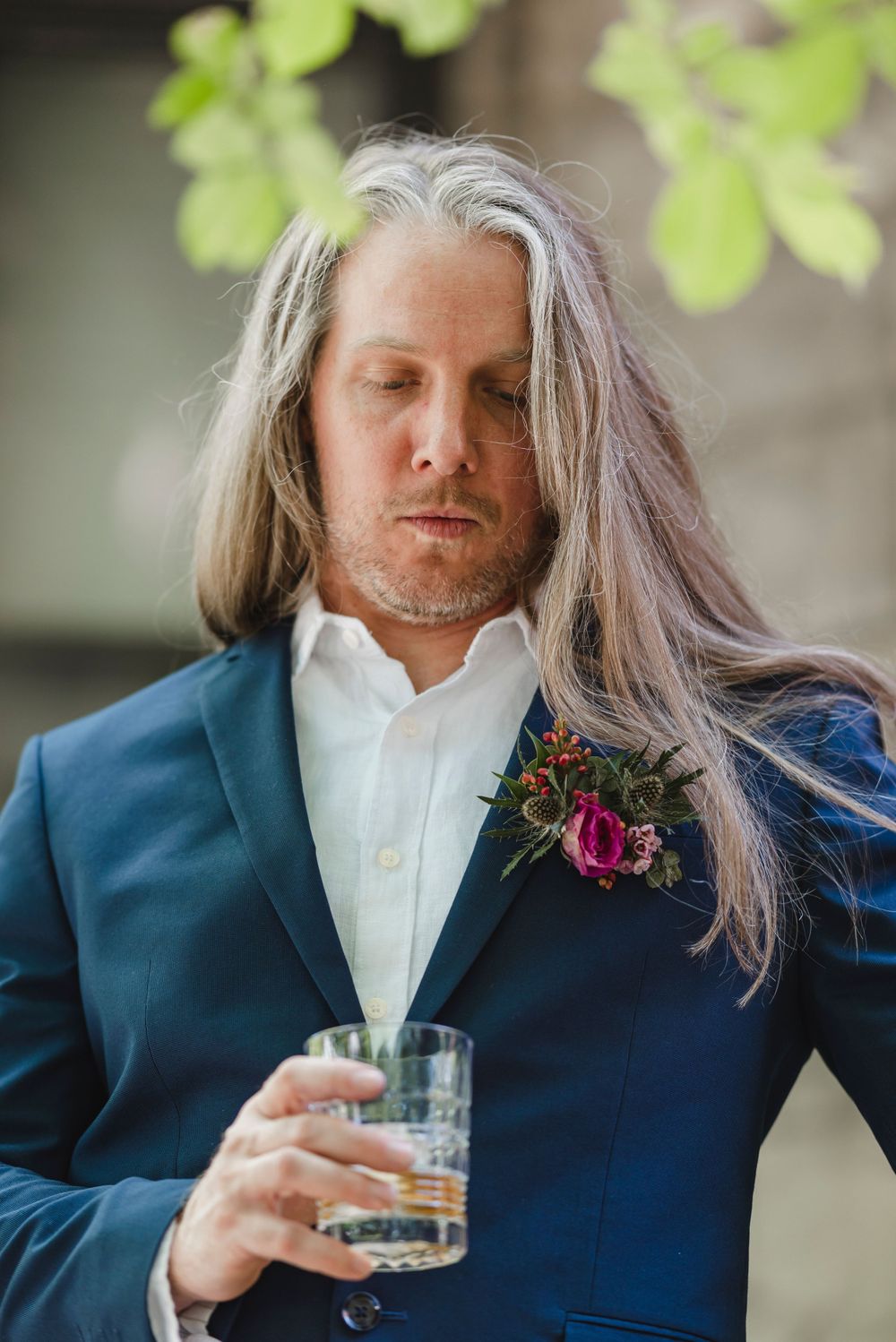 Person in navy suit with boutonniere and long hair enjoying a drink.