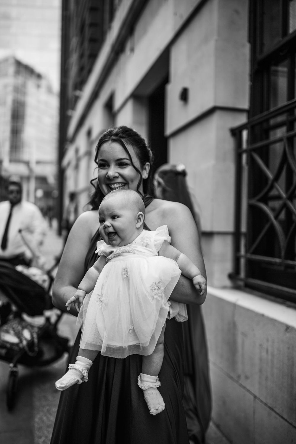 A parent holds a joyful baby wearing a white dress against an urban backdrop.